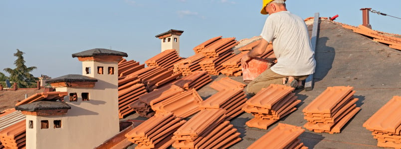roofing: construction worker on a roof covering it with tiles - roof renovation: installation of tar paper, new tiles and chimney